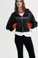 Sudadera negra con capucha, cierre frontal de cremallera metálica y mangas largas con puños. Presenta un diseño corto con bolsillo tipo canguro y detalle de texto 'California' aplicado con brillos en el frente.