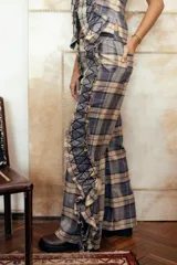 Pantalón cuadrillé de tiro medio con estampa a cuadros en tonos beige, gris y azul. Presenta diseño de perneras anchas, bolsillos laterales y traseros, y detalles de volados con costuras decorativas en zig-zag a lo largo de las piernas.