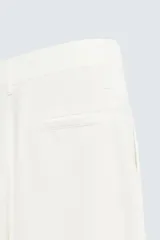 Pantalón blanco de corte relajado (relaxed fit) con pliegues frontales, bolsillos laterales y bolsillos traseros de vivo. Cierre frontal con cremallera y botón.