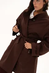 Trench color chocolate con cuello alto, cierre frontal con botones y cinturón ajustable en la cintura. Presenta mangas largas con puños ajustables mediante tiras y hebillas, y bolsillos frontales.
