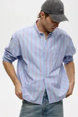 Camisa de corte relajado confeccionada en algodón, con diseño de rayas verticales en tonos azul, blanco y rojo. Presenta cuello americano, cierre frontal con botones, bolsillo en el pecho y mangas largas con puños abotonados.