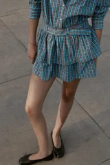 Vestido corto de manga larga con cuello solapa y cierre frontal de botones a presión. Presenta un diseño a cuadros en tonos azul y marrón, cintura ajustable con cinturón de hebilla y falda con volantes en el bajo.