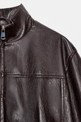 Chaqueta estilo bomber corta de efecto piel en color marrón oscuro, con cuello subido, cierre frontal con cremallera y cintura elástica fruncida.