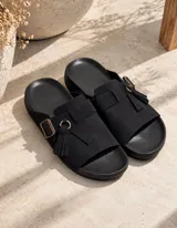 Sandalias planas color beige, fabricadas en gamuza, con tira ancha en el empeine, hebilla metálica de ajuste y detalle de flecos colgantes con argolla.