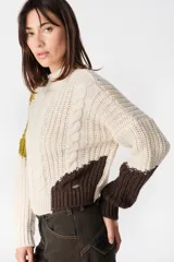 Sweater de punto con diseño de bloques de color en tonos crema y marrón. Presenta cuello redondo, mangas largas y una silueta holgada con detalle de placa metálica decorativa en el lateral.