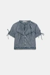 Camisa de cuello redondo con lazada frontal, manga corta abullonada con lazos ajustables y cierre frontal con botones. Presenta detalles de bordados y encaje a tono sobre una base de tela tipo chambray.