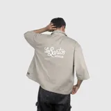Sobre camisa de gabardina color beige, con corte boxy fit, cuello clásico, cierre frontal con botones y etiqueta decorativa en el ruedo inferior.