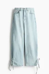 Jean de tiro alto confeccionado en denim de algodón rígido, con diseño curvo y voluminoso en las piernas, tipo barrel. Presenta cinco bolsillos, cierre frontal con botón y detalles de tiras para anudar en los tobillos.