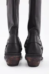 Bota de caña alta estilo western confeccionada en cuero vacuno negro. Presenta un diseño robusto con taco cuadrado de madera, punta redondeada y una tira decorativa con herrajes metálicos y tachas en la zona del tobillo.