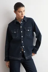 Camisa de jean azul oscuro con cierre frontal de botones metálicos a presión y dos bolsillos delanteros con solapa en el pecho. Presenta mangas largas con puños anchos doblados y cuello camisero clásico.