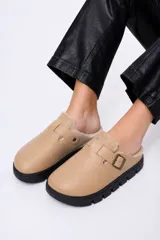 Zueco de cuero sintético color beige, con interior forrado en corderito sintético, tira ajustable con hebilla metálica y suela de goma negra con plataforma dentada.