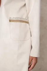 Chaqueta larga color beige con diseño abierto, cuello redondo y mangas largas. Presenta detalles bordados artesanales con aplicaciones de cuentas y toques dorados en la zona de los hombros.