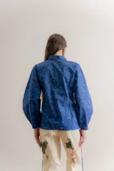 Camisa de popelina azul con estampado toile de jouy, cuello clásico y cierre frontal con botones. Presenta un diseño ajustado en la cintura mediante cordones laterales estilo corset y mangas largas abullonadas.