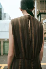 Vestido midi sin mangas de cuello redondo, con diseño de rayas verticales en tonos marrones. Presenta un detalle drapeado en el frente, volantes laterales y un bajo asimétrico. Cierre posterior con cremallera oculta.