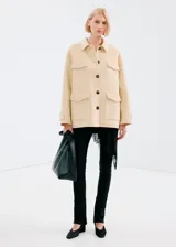 Chaqueta de corte recto color beige, con cuello camisero, cierre frontal de botones oscuros y cuatro bolsillos con solapa en el frente.