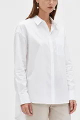 Camisa blanca de algodón, de corte holgado y largo tipo túnica, con cuello camisero, cierre frontal con botones y un bolsillo aplicado en el pecho.