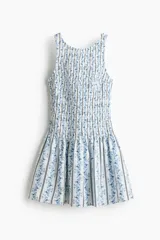 Vestido corto de algodón con sección superior fruncida y pollera con vuelo, sin mangas, con escote redondo al frente y en U en la espalda, con estampado combinado de rayas y motivos florales en tonos azules sobre fondo blanco.