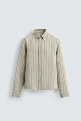 Camisa de algodón de corte relajado, con cuello solapa abotonado (button-down) y manga larga terminada en puño con botón. Presenta un patrón de rayas verticales finas en tonos beige claro y marrón/verde oliva.