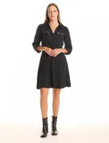 Vestido corto negro de crepe, con cuello solapa y escote en V. Presenta bolsillos simulados con cierres metálicos dorados en la parte delantera y mangas largas regulables con presilla y botón. La cintura es elastizada.