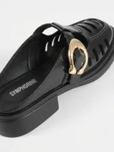 Zuecos negros de charol estilo mocasín destalonado, con perforaciones laterales tipo huarache, tacón bajo cuadrado y hebilla metálica dorada decorativa en el empeine.