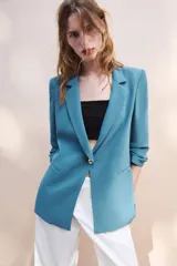 Blazer negro de cuello con solapas de muesca y hombreras, con mangas tres cuartos drapeadas, bolsillos de vivo simulados y cierre frontal con un botón dorado. Forro interior a tono.