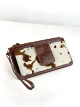 Billetera tipo porta celular de cuero, con diseño combinado en cuero liso marrón y cuero con pelo vacuno estampado. Cuenta con cierre superior, correa de muñeca y solapa frontal con cierre magnético y logo de búho grabado.