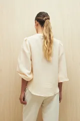 Camisa color crudo a rayas verticales finas, de cuello mao y mangas 3/4 abullonadas con elástico en los puños. Cierre frontal con botones de nácar.