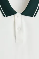 Polo de manga corta en piqué de algodón, color blanco con cuello y bordes de mangas en color verde oscuro con detalles de líneas blancas. Presenta tapeta con botones.