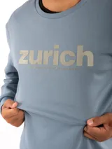 Buzo cerrado color celeste, de manga larga, con estampa frontal con la palabra "zurich" y texto complementario.