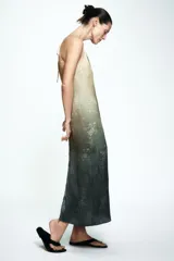 Vestido midi de estilo lencero con escote en pico y tirantes finos. Presenta un diseño degradado que va de un tono beige claro a un verde oscuro, con estampado floral sutil. Incluye lazada en la espalda y abertura en el bajo.