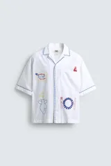 Camisa blanca de algodón con cuello bowling y manga corta, corte relajado. Presenta bordados a contraste en azul, rojo y amarillo que simulan elementos de cocina y artesanía, además de ribetes de pespunte azul en cuello, puños y abertura frontal.