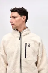 Campera de sherpa con diseño capitoneado en color crema. Presenta cierre frontal completo, cuello alto, bolsillos laterales y un bolsillo en el pecho con cierre en contraste. Incluye detalles en nylon y bordado decorativo en el pecho.
