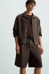 Bermuda de corte regular fit, color marrón oscuro, confeccionada en tejido con lino. Presenta pliegues frontales, bolsillos delanteros y bolsillos traseros de vivo. Cierre frontal con cremallera y botón.