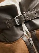 Campera estilo aviador de color marrón, confeccionada en tejido tipo gamuza con forro interno y detalles de corderito sintético en cuello y ruedo. Presenta cierre frontal con cremallera, bolsillos laterales inclinados y detalles en cuero sintético oscuro en las costuras y solapas.