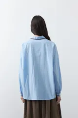 Camisa blanca de manga larga con detalles bordados en contraste a lo largo de la botonadura, el cuello y los puños. Presenta una silueta relajada y un diseño clásico con un toque artesanal.