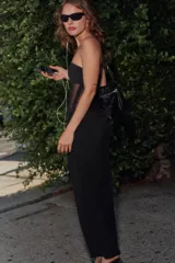 Vestido midi de escote strapless y hombros descubiertos, confeccionado en tejido negro combinado con detalle drapeado en la parte frontal de la falda. Presenta un diseño entallado y cierre lateral con cremallera oculta.
