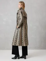 Trench de ecocuero con estampado animal print de serpiente en tonos beige, marrón y negro. Presenta cuello con solapa ancha, doble abotonadura frontal con botones negros, cinturón ajustable con hebilla metálica, mangas largas con presillas y botones en los puños, canesú suelto en la espalda, bolsillos laterales y forro interno.