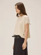 Remera tejida color beige de corte relajado, con mangas cortas y detalle de costuras frontales marcadas. Incluye un pequeño bordado distintivo en el borde inferior.