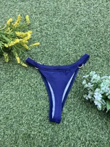 Bikini azul oscuro con tela texturada. El corpiño es triangular con una argolla de zamak en el centro y tirantes finos. La bombacha es colaless con argollas metálicas en los laterales.