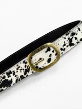Cinto de cuero de pelo con estampado animal print tipo vaca en color blanco y negro, con hebilla metálica ovalada en tono bronce viejo con la marca grabada.