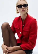 Chaqueta de tweed color rojo con cuello redondo, manga larga y diseño corto. Presenta cuatro bolsillos de parche en la parte frontal, cierre de botones metálicos y terminaciones deshilachadas.