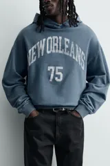 Sudadera tipo hoodie de corte relajado en color azul con efecto lavado. Presenta capucha, mangas largas, puños y ruedo en rib, con estampado frontal en color blanco con texto y números.