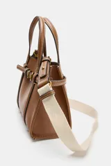 Bolso tipo mini shopper de cuero sintético color marrón caramelo, con costuras blancas visibles. Posee asas de mano y una correa bandolera ancha de tela color beige. El frente tiene un detalle de tira con hebilla metálica dorada.