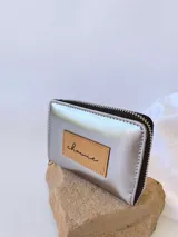Billetera pequeña de cuero color plateado con acabado metalizado, cierre perimetral metálico y parche de cuero en tono natural con el logo de la marca grabado.