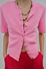 Chaqueta de punto de manga corta en color rosado, con cuello redondo y cierre frontal de botones dorados.