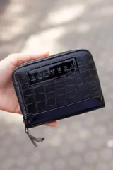 Billetera chica de cuero negro con textura tipo cocodrilo y cierre de cremallera. Tiene el logo de la marca Austera grabado en la parte superior.