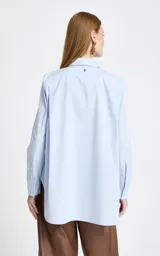 Camisa de algodón color celeste con cuello clásico, manga larga con puños y cierre frontal de botones. Presenta apliques decorativos de pedrería brillante distribuidos por toda la prenda.