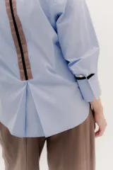 Camisa de manga larga color celeste claro, con cuello camisero y botones frontales. Presenta un detalle de cinta negra alrededor del puño doblado.