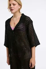 Vestido corto de tejido calado en color negro, con cuello polo, cierre frontal parcial con botones y mangas cortas. Presenta un diseño holgado y textura abierta.