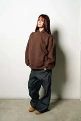 Hoodie oversize marrón oscuro con capucha y bolsillo canguro frontal. Presenta un logo bordado en tono gris claro en el pecho.